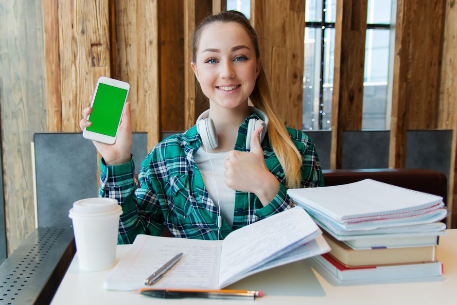 Smiling young woman using a smartphone while studying with books, showcasing modern education.